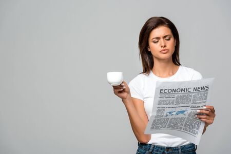 beautiful confused woman holding coffee cup while reading economic newspaper, isolated on greyの写真素材