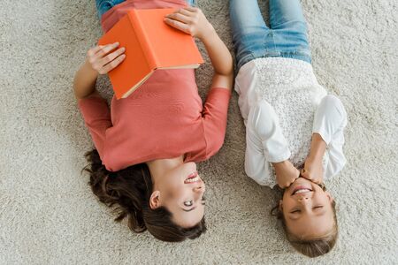 top view of happy babysitter holding book while looking at kid lying on carpetの写真素材