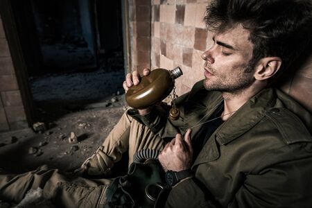 handsome man holding flask while sitting in abandoned building, post apocalyptic concept