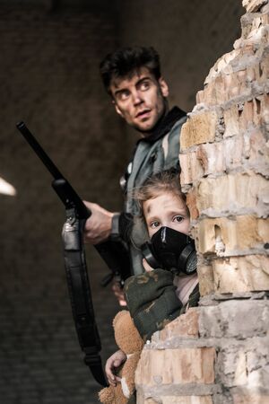 selective focus of kid in gas mask near handsome man with gun, post apocalyptic concept
