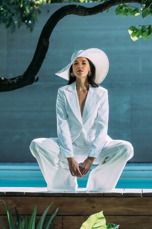 attractive woman in white suit and hat posing and looking at camera outsideの写真素材
