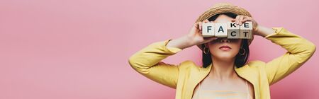 panoramic shot of asian woman holding wooden cubes in front of face with fake and fact lettering isolated on pinkの写真素材