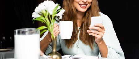 cropped view of smiling woman with cup of coffee using smartphone in cafe