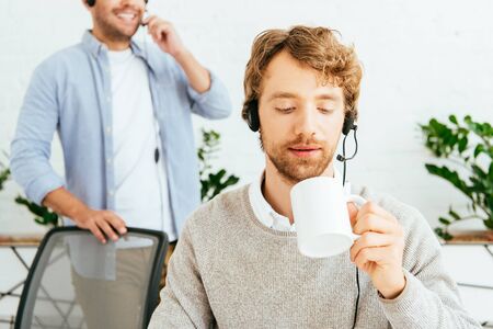 selective focus of handsome bearded broker holding cup near coworker in officeの写真素材