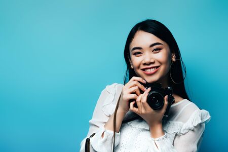 happy asian woman in white blouse holding digital camera on blue backgroundの写真素材
