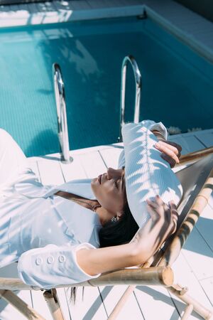 high angle view of attractive woman in white suit and hat sitting on deck chair outsideの写真素材