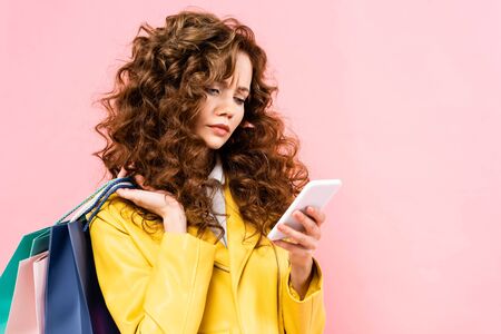 beautiful thoughtful girl using smartphone and holding shopping bags, isolated on pink