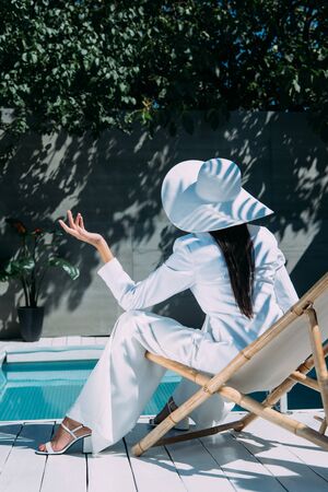 back view of woman in white suit and hat sitting on deck chair outsideの写真素材