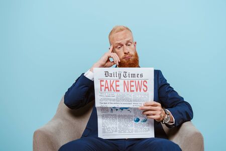 thoughtful bearded businessman reading newspaper with fake news while sitting in armchair, isolated on blueの写真素材
