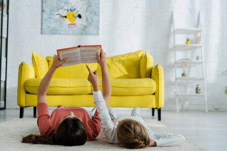 child pointing with finger at book while lying on carpet with babysitter in living roomの写真素材