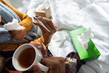 cropped view of sick couple holding thermometer and hot drink in bed with napkinsの写真素材