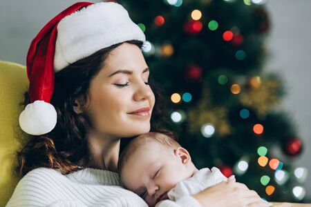 happy woman in santa hat sitting with closed eyes and holding sleeping babyの写真素材