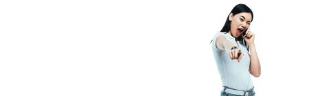 excited brunette asian woman talking on smartphone and pointing with finger at camera isolated on...