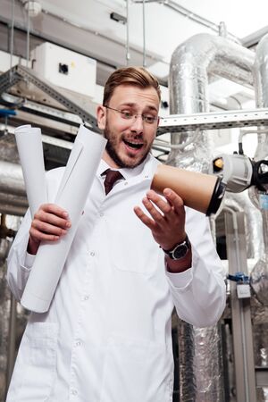 worried engineer in white coat and glasses holding blueprints and looking at falling paper cup near...