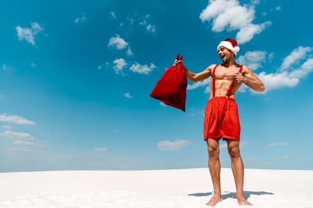 handsome and sexy man in santa hat pointing with finger at santa sack on beach in Maldivesの写真素材