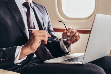cropped view of businessman in suit with laptop holding glasses in private planeの写真素材