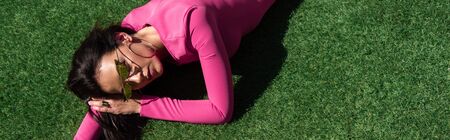 panoramic shot of attractive woman in dress and sunglasses posing and lying on grass outside