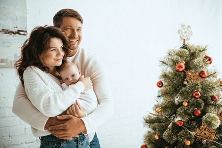 happy man embracing wife with baby while standing near christmas treeの写真素材