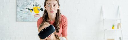 panoramic shot of babysitter looking at camera near kid in virtual reality headset