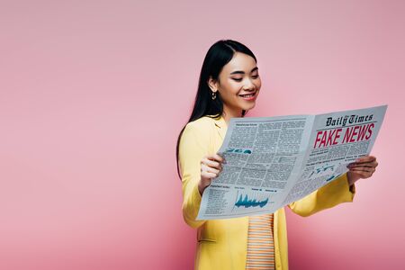 happy asian woman in yellow outfit reading newspaper with fake news isolated on pinkの写真素材