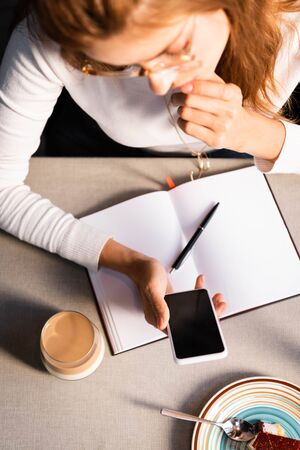overhead view of woman with notepad using smartphone in cafe with glass of coffeeの写真素材