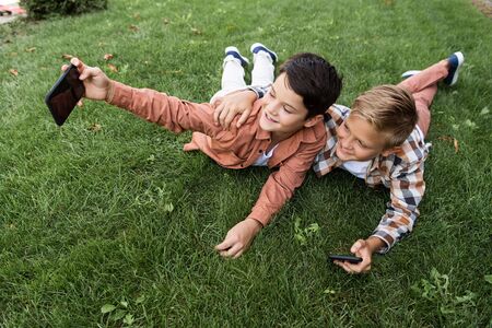cheerful boy taking selfie on smartphone while lying on grass with brotherの写真素材
