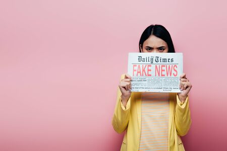 sad asian woman in yellow outfit holding newspaper with fake news isolated on pinkの写真素材