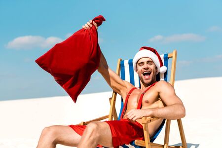 handsome and sexy man in santa hat holding santa sack and sitting on deck chair on beach in Maldivesの写真素材