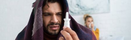 sick man holding nasal spray in bedroom with woman behind