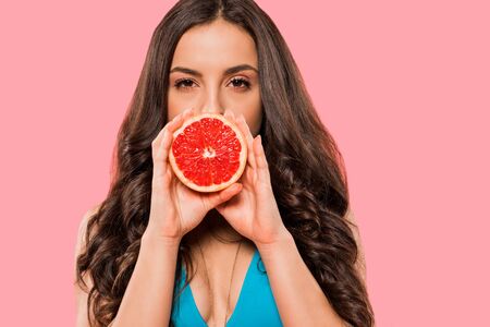 young woman in swimsuit covering face with half of grapefruit isolated on pinkの写真素材