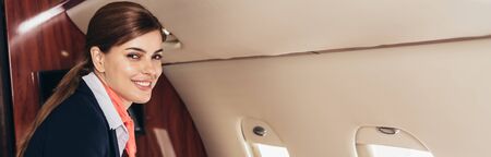 panoramic shot of smiling flight attendant in uniform looking at camera in private plane