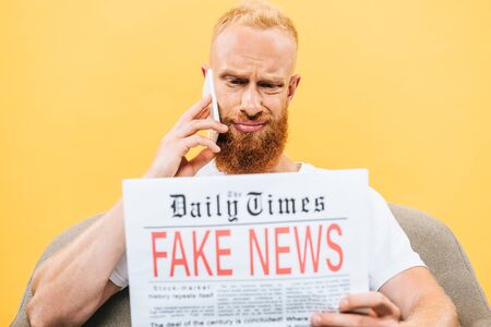 skeptical man reading newspaper with fake news and talking on smartphone while sitting on armchair, isolated on yellowの写真素材