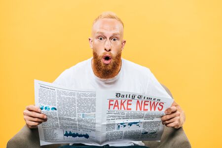 surprised man reading newspaper with fake news while sitting on armchair, isolated on yellowの写真素材