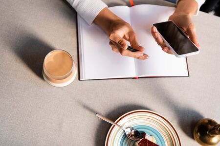 partial view of woman with notepad using smartphone in cafe with cake and coffeeの写真素材
