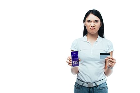 displeased brunette asian girl holding credit card and smartphone with healthcare app isolated on whiteの写真素材