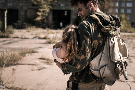 handsome man with backpack holding in arms child outside, post apocalyptic concept