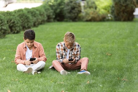 two concentrated brothers sitting on green lawn and using smartphones
