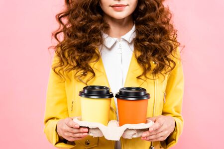 cropped view of curly woman holding paper cups with coffee, isolated on pinkの写真素材