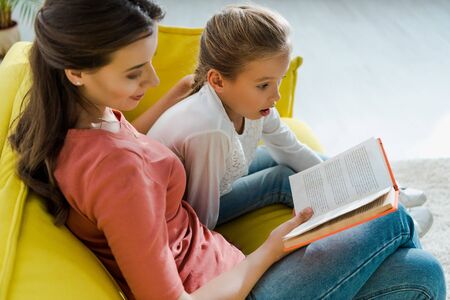 selective focus of surprised kid sitting on sofa with happy babysitter reading book