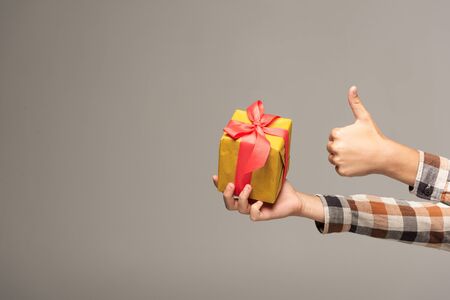 cropped view of boy holding yellow gift box and showing thumb up isolated on greyの写真素材