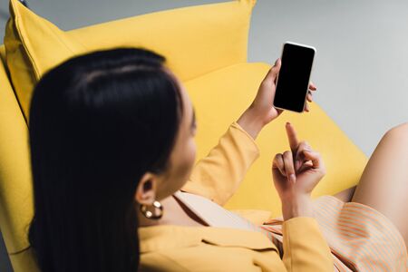 brunette businesswoman sitting on yellow sofa and showing middle finger to smartphone with blank screen isolated on greyの写真素材