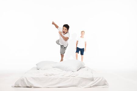 excited boy levitating over bed while smiling brother standing on white backgroundの写真素材