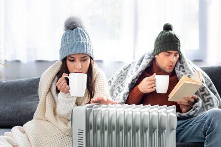 attractive girlfriend with cup of tea and boyfriend reading book and warming up near heater