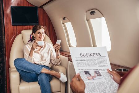 selective focus of boyfriend reading newspaper and girlfriend holding cup and smartphone in private planeの写真素材