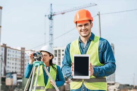 Smiling surveyor holding digital tablet with blank screen and colleague with measuring level at...