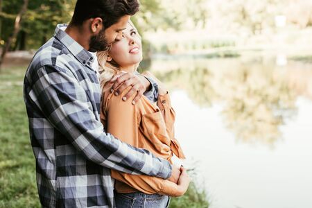handsome man embracing happy girlfriend while standing near lake in parkの写真素材