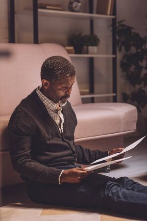 discouraged african american man sitting on floor and holding utility bills