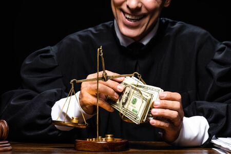 cropped view of smiling judge in judicial robe sitting at table and holding dollar banknotes isolated on blackの写真素材