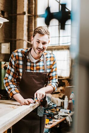 selective focus of cheerful woodworker holding chisel while carving wood in workshopの写真素材