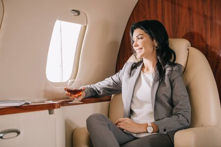 cheerful businesswoman in formal wear holding glass with cognac in private jetの写真素材